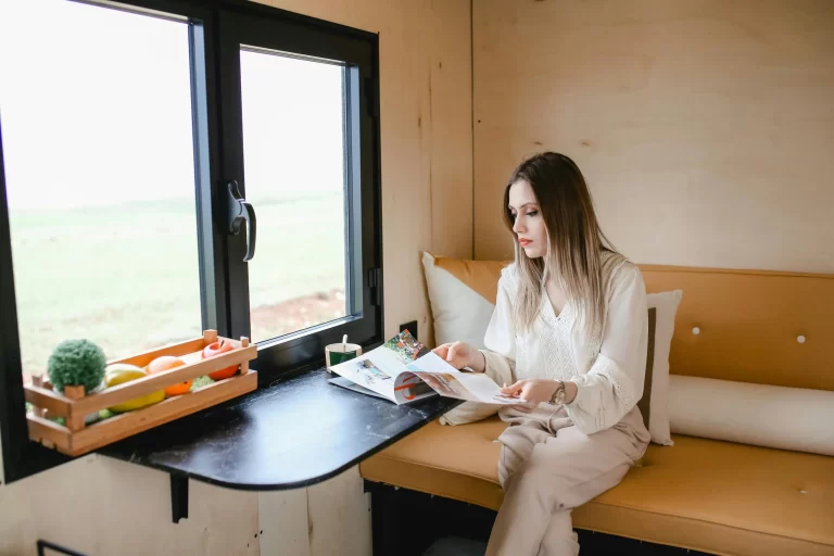 Woman reading magazine in a cozy tiny house with a scenic view.